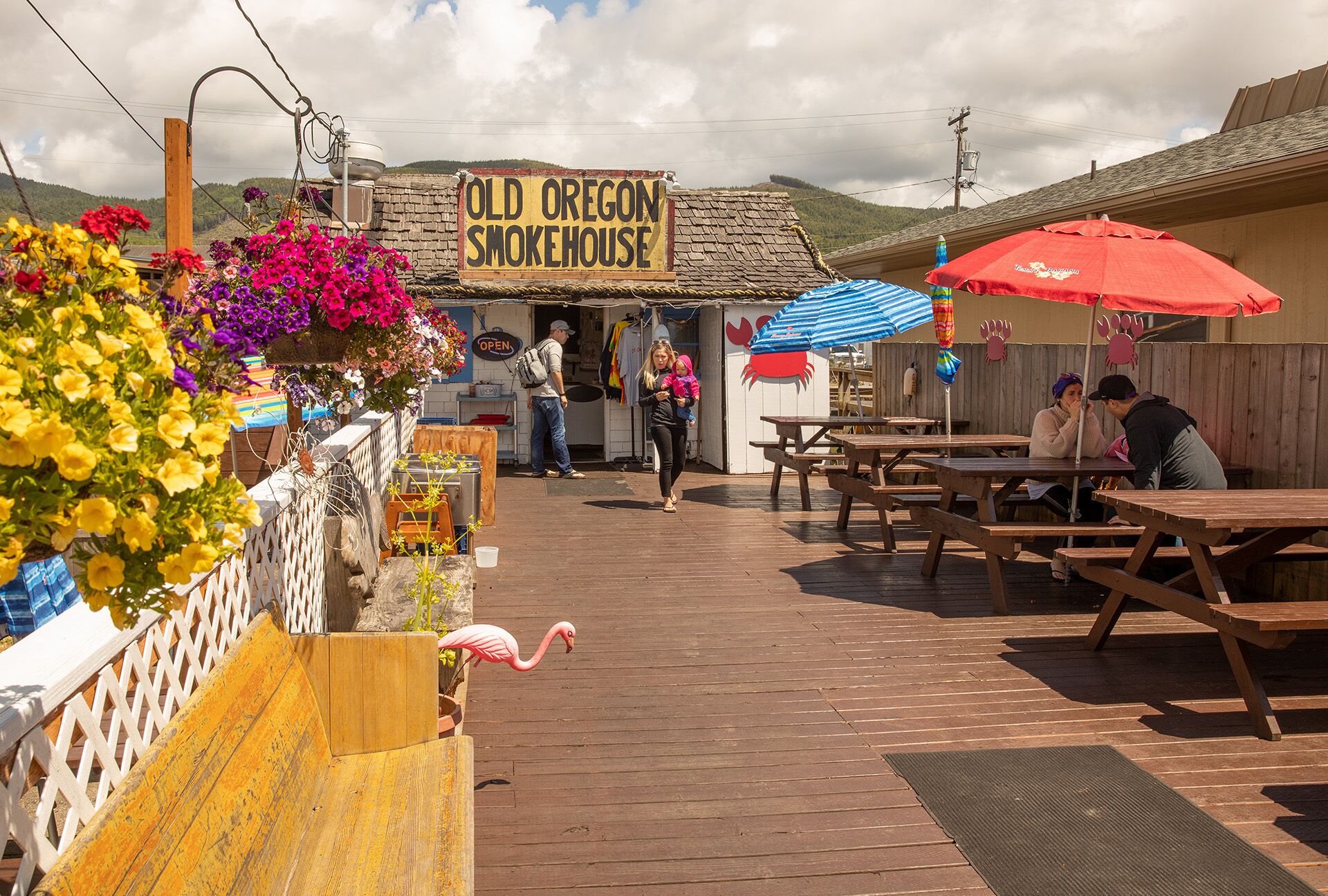 An outdoor seating area with wooden tables and benches, a sign reading "Old Oregon Smokehouse," and a wooden fence with colorful flowers.