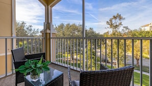 A balcony with wicker furniture and a glass table, overlooking a residential area with trees and a clear sky.