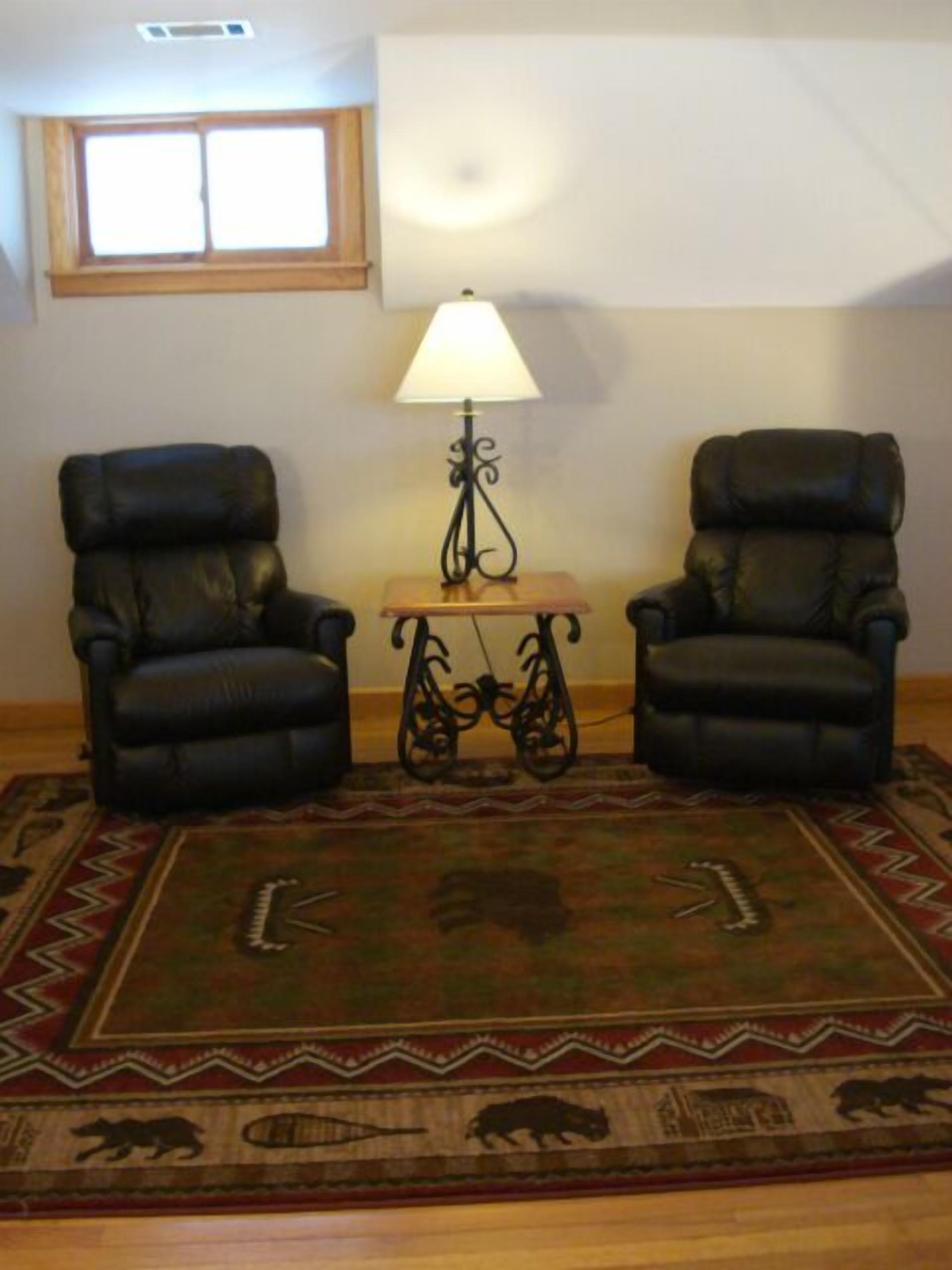 A room with two black leather chairs, a small wooden table with a lamp, and a patterned rug.