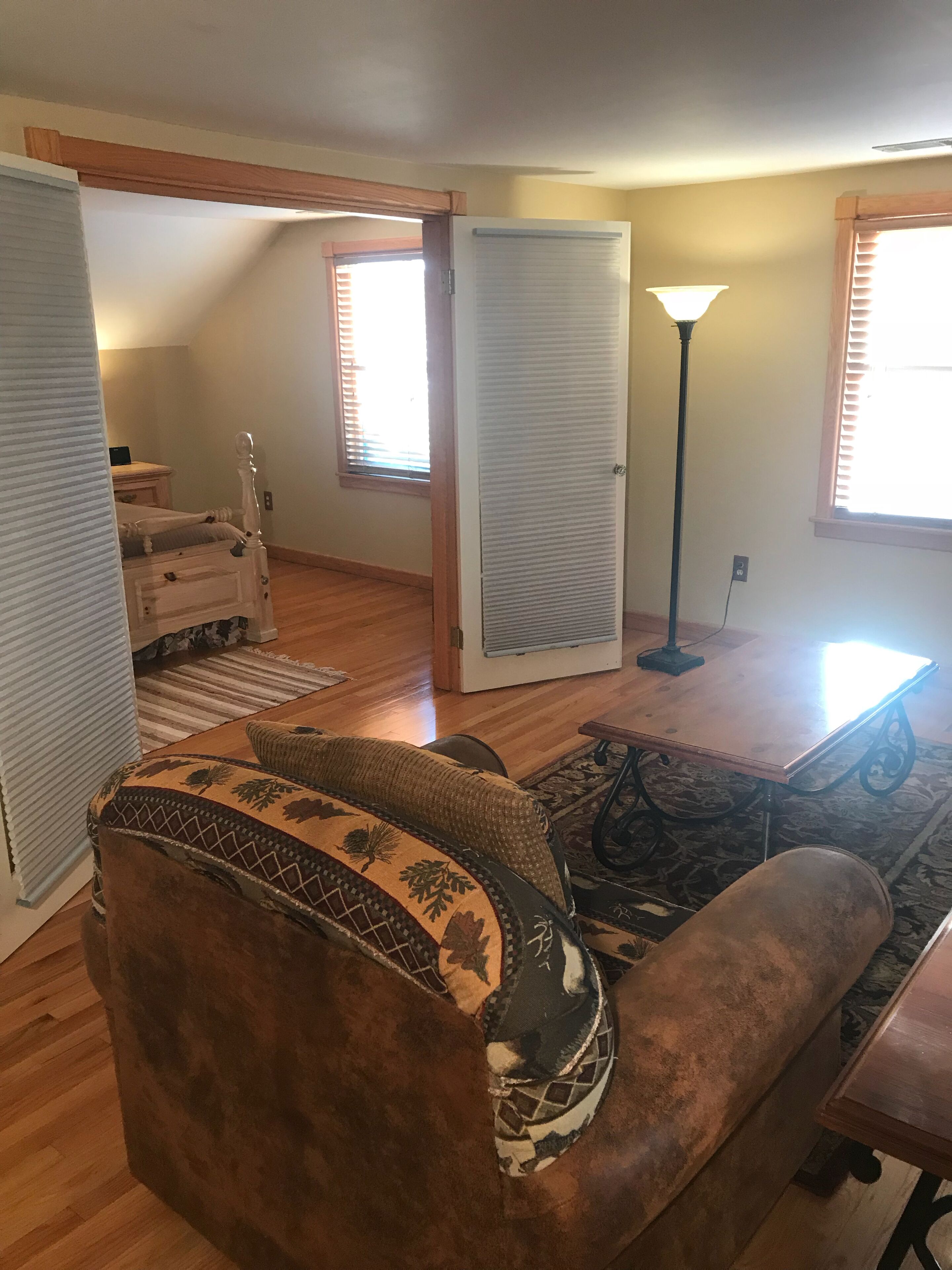 A living room with a brown sofa, a patterned rug, a wooden coffee table, and a lamp. There is a door leading to another room, and a window with blinds.