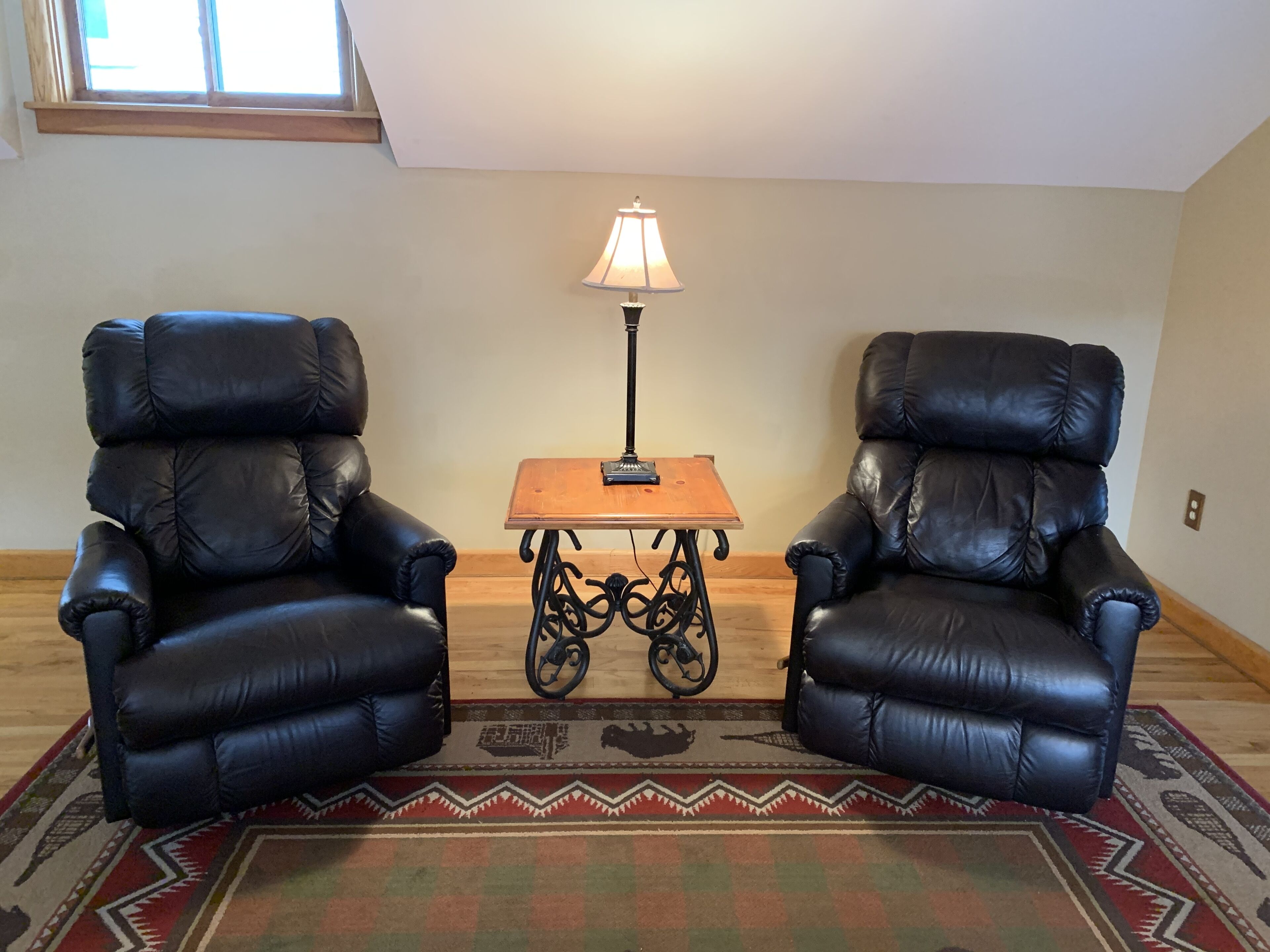 A room with two black leather armchairs, a small wooden table with a lamp, and a patterned rug.