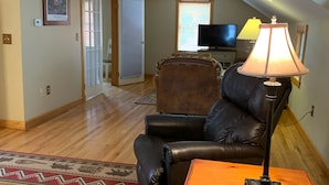 A spacious living room with a leather sofa, a wooden coffee table, and a lamp. A television is mounted on the wall, and a door leads to another room. - Great Location Next to Fishtown in Leland (Leland)