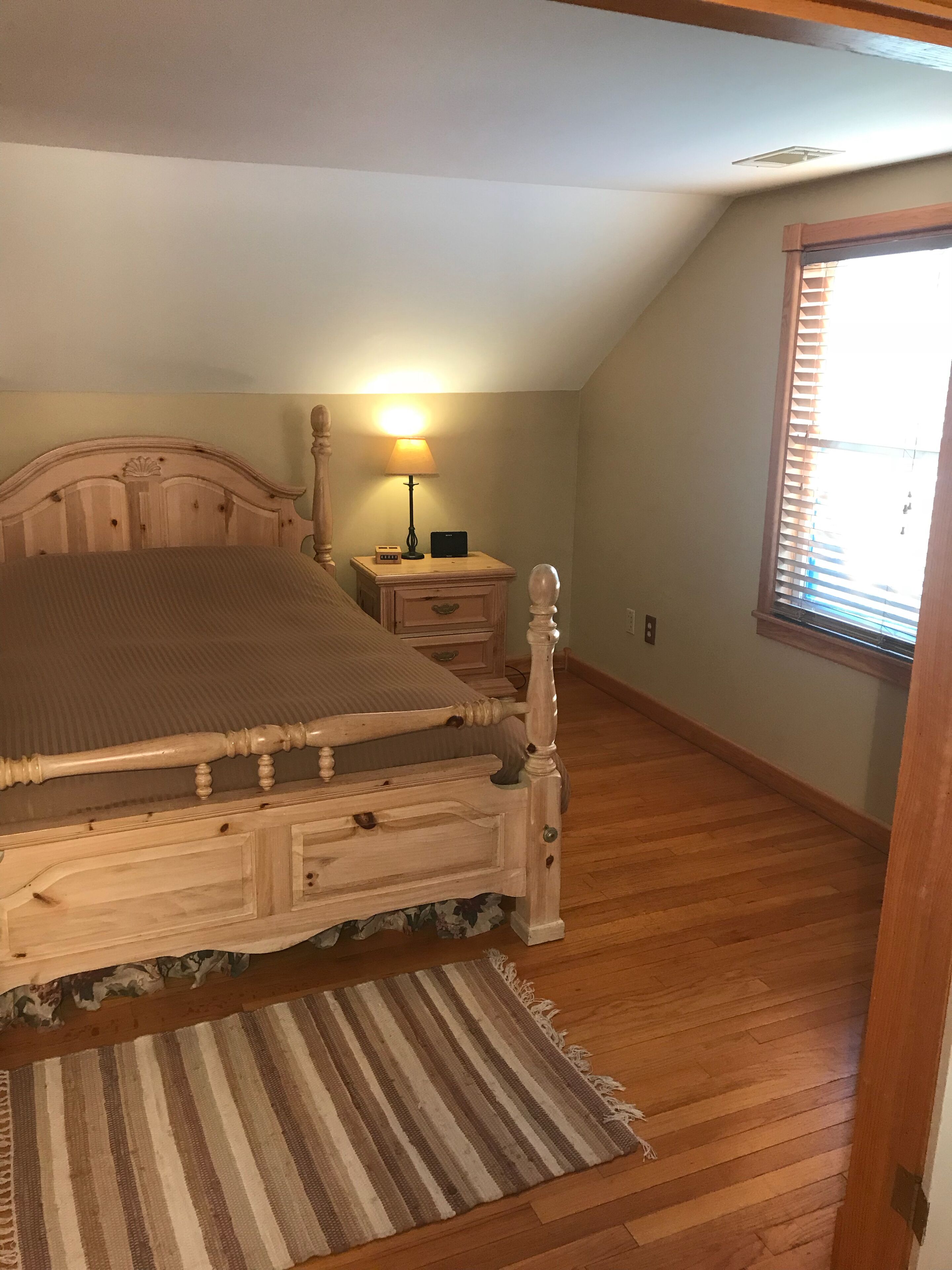 A bedroom with a wooden bed, a nightstand, a lamp, and a window with blinds.