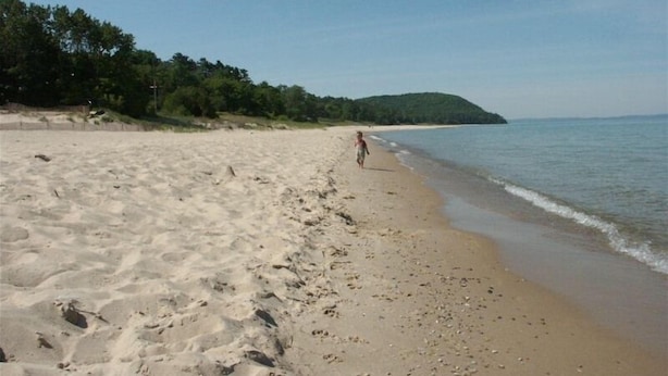 A sandy beach with footprints leading to the water, a person walking, and a forested hill in the background. - Great Location Next to Fishtown in Leland (Leland)