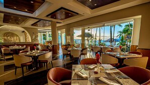 A spacious restaurant with multiple tables and chairs, large windows offering a view of the ocean and palm trees, and a modern interior design with marble accents. - Huge true Beachfront condo w/ full resort amenities & washer/dryer (Playa del Carmen)