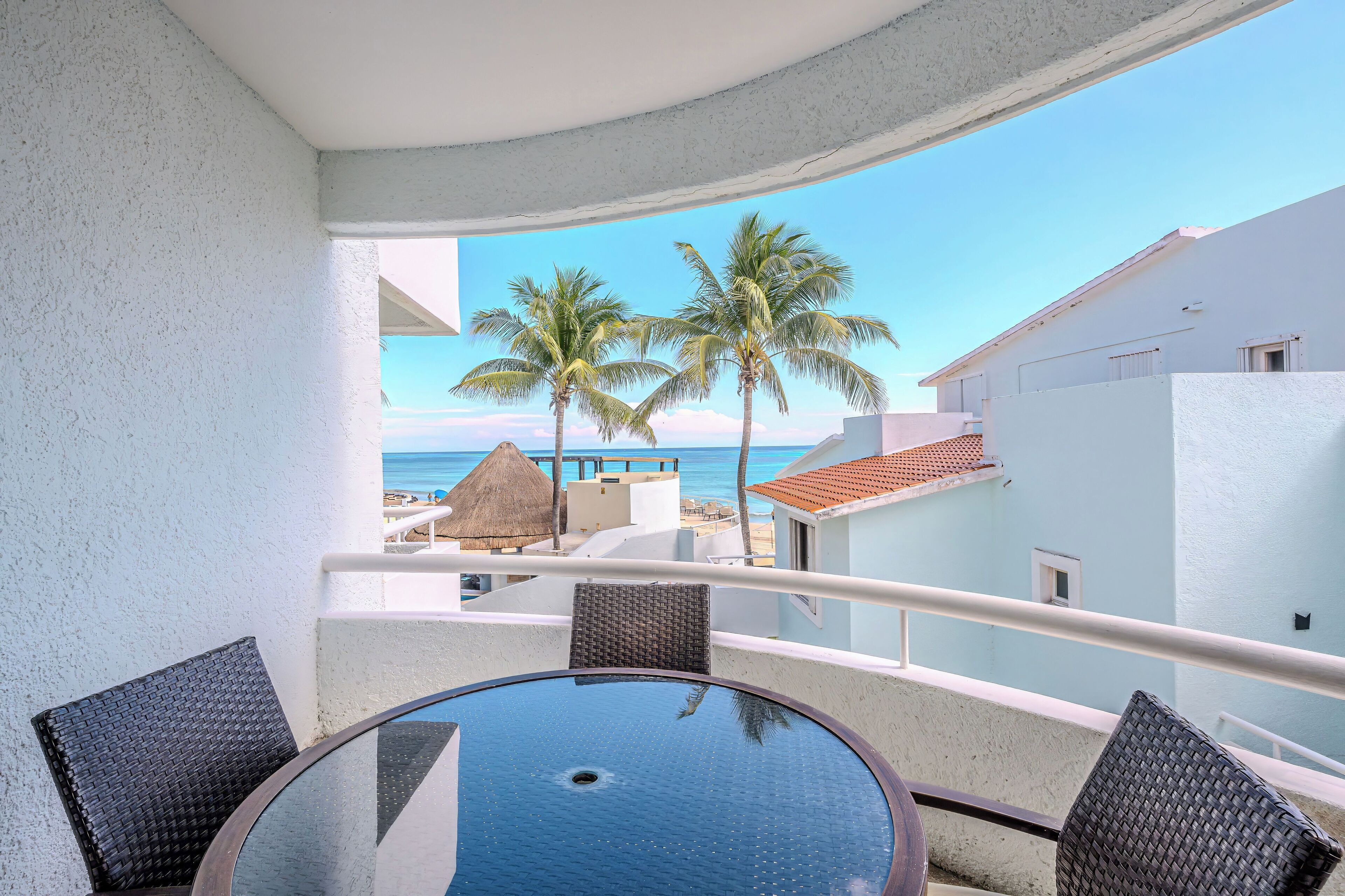 Ein Balkon mit rundem Tisch und Stühlen, der Blick auf einen Strand mit Palmen und einem Gebäude mit Reetdach gewährt.