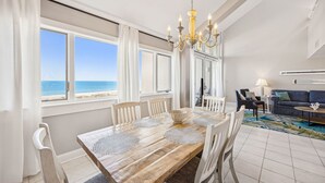 Ein Essbereich mit Holztisch, weißen Stühlen und einer Kronleuchter. Große Fenster bieten Blick auf den Ozean. Im Hintergrund sind eine Couch und eine Lampe zu sehen.