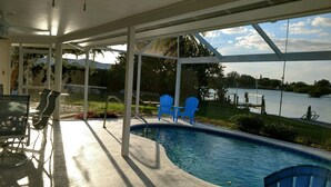 A swimming pool with a view of a lake, surrounded by a covered patio area with chairs and a ceiling fan.