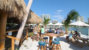 A beachside restaurant with outdoor seating, palm trees, and a view of the water and buildings in the background.