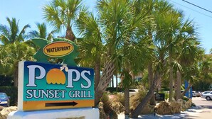 A sign for 'Pops Sunset Grill' with a 'Waterfront' label, surrounded by palm trees and rocks.