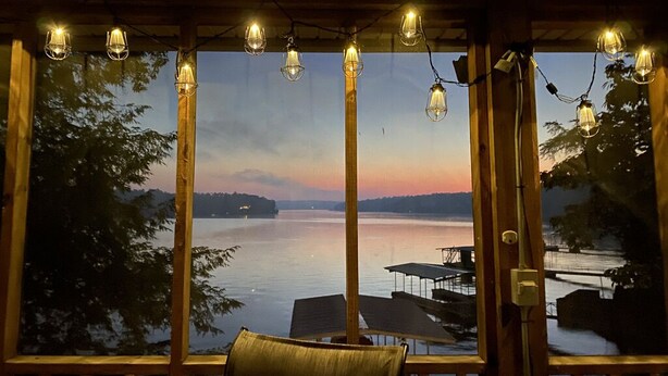 A wooden deck with a view of a lake and distant mountains, string lights, and a chair.