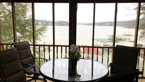 A room with a round glass table and two chairs, a potted plant, and a view of a body of water through large windows.