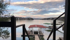 A wooden deck leading to a dock with a covered seating area, overlooking a calm lake with distant land and cloudy sky.