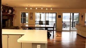 A spacious living area with a wooden floor, a dining table, and a fireplace. There are large windows offering a view of the outdoors.