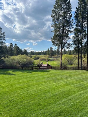 A well-maintained grassy field with a wooden fence, tall trees, and a distant view of a cabin. - RIVER FRONT front property with Hot Tub! Bring the dogs! (Bend)
