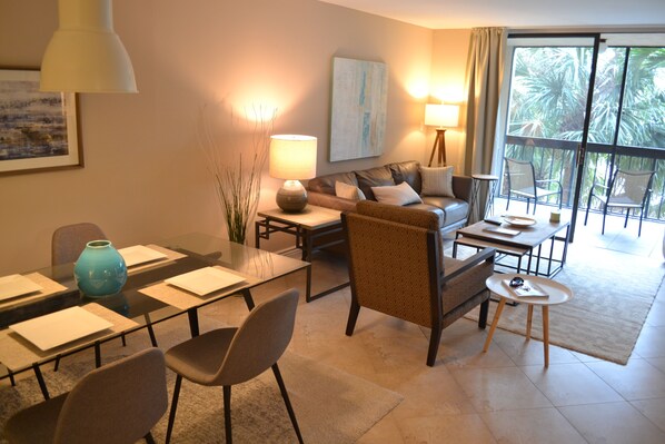 A modern living room with a glass dining table, comfortable seating, a sofa, and a view of the outdoors.