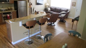 Una cocina moderna con una isla central, taburetes de bar, una mesa de comedor y una sala de estar con un sofá y televisor.