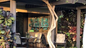 Eine Terrassenfläche mit Außenmöbeln, einem kleinen Tisch und Stühlen. In der Mitte steht ein Baumstamm, und rund um die Sitzgruppe sind verschiedene Pflanzen verteilt.