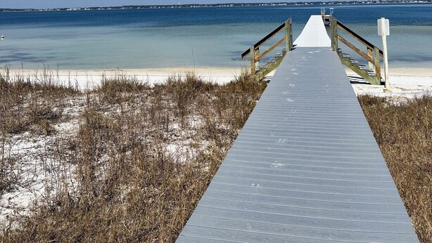 Ein hölzerner Steg führt zu einem Strand mit klarem, blauem Wasser und einer entfernten Küstenlinie.