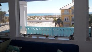 Ein Zimmer mit großem Fenster mit Blick auf einen Strand, ein Balkon mit verzierten Geländern und eine Ansicht eines gelben Gebäudes gegenüber der Straße.