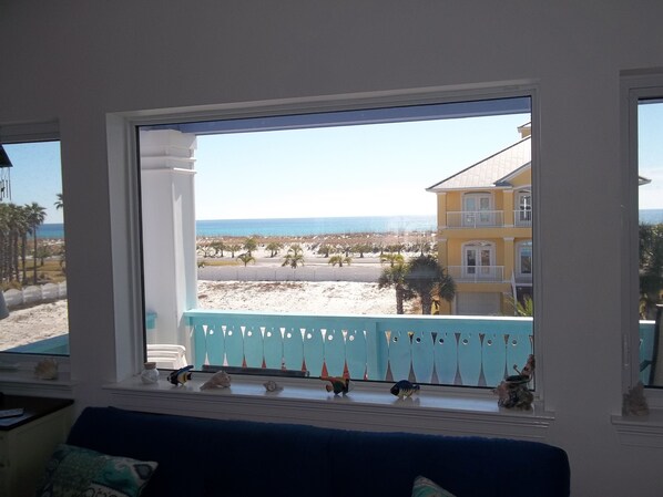 A room with a large window overlooking a beach, a balcony with decorative railings, and a view of a yellow building across the street.