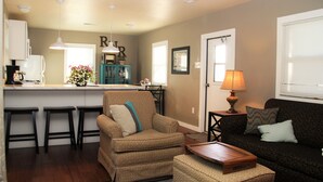 A living room with a sofa, armchair, ottoman, and a coffee table. There is a kitchen area with a refrigerator, sink, and bar stools. A lamp and a picture frame are on the wall. - Rest, Relaxation, & Quick Access to Ohiopyle Await (Ohiopyle)