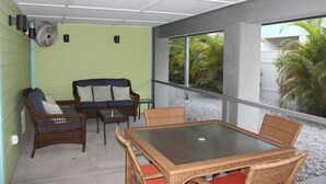 Uma área de varanda coberta com móveis de vime, uma mesa de vidro e vista para palmeiras através de grandes janelas.