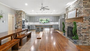 A spacious kitchen with a central island, wooden benches, a brick fireplace, and a ceiling fan. - Secluded Country Retreat with Boutique Hotel Vibes plus Pool/Hot Tub sleeps 50+ (Crawford)