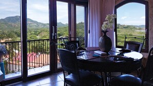 Un balcón con una mesa y sillas, un florero con flores, y vistas a colinas verdes.