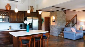Una cocina con gabinetes de madera, una isla central con taburetes de bar, un refrigerador y lámparas colgantes.