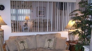A living room with a sofa, a coffee table, and a window with vertical blinds. - Pass-a-Grille Sunsets Are the Best! (st. pete Beach)