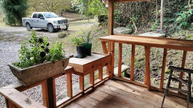 A wooden deck with a bench and a table, surrounded by potted plants and a view of a white pickup truck in the background.