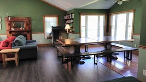 A spacious living room with a large wooden table and benches, a fireplace, a bookshelf, and a large window with curtains.