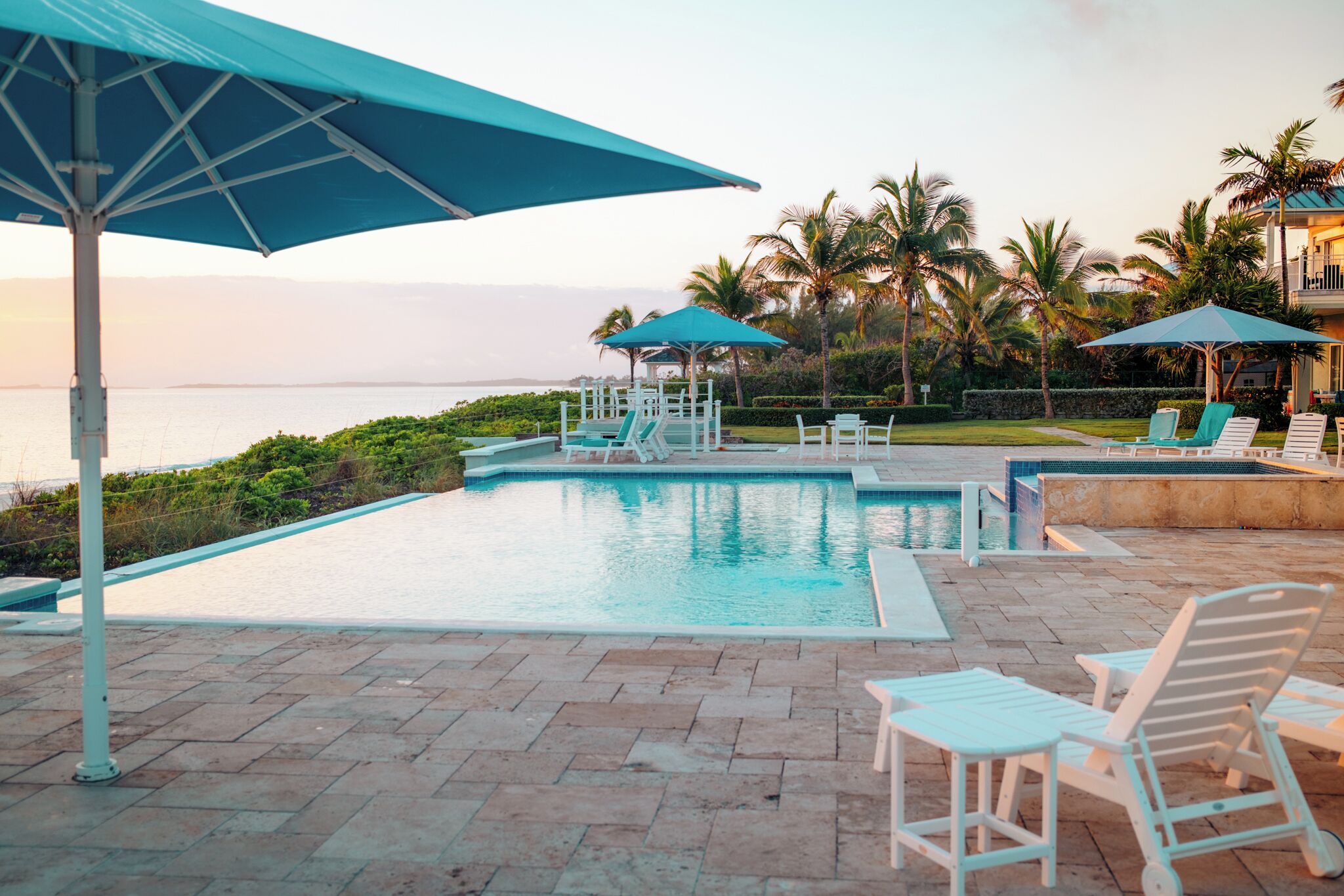 A pool area with lounge chairs and umbrellas, surrounded by palm trees and a view of the ocean.