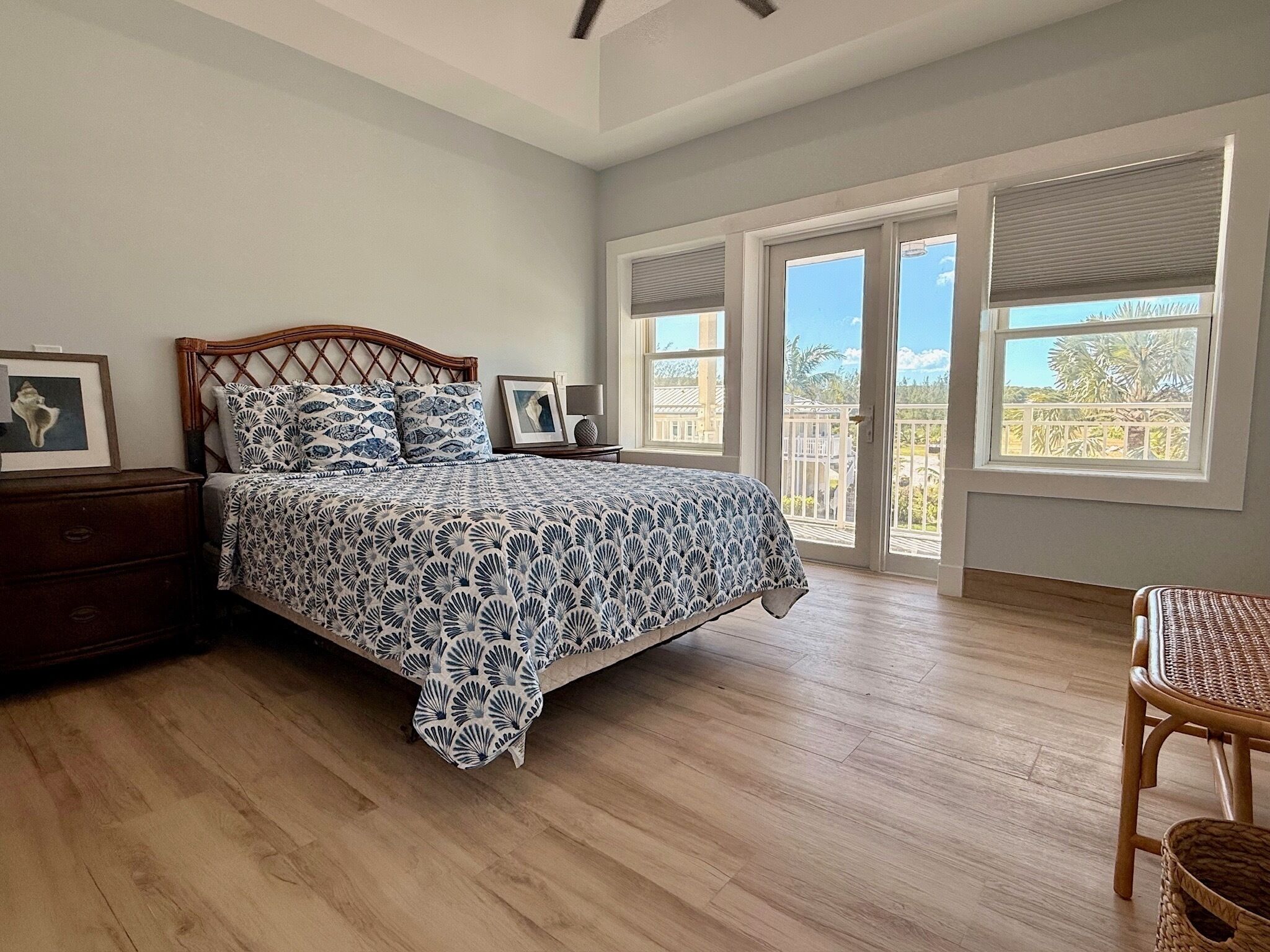 A bedroom with a bed, wooden nightstands, a wicker chair, and a view through large windows.