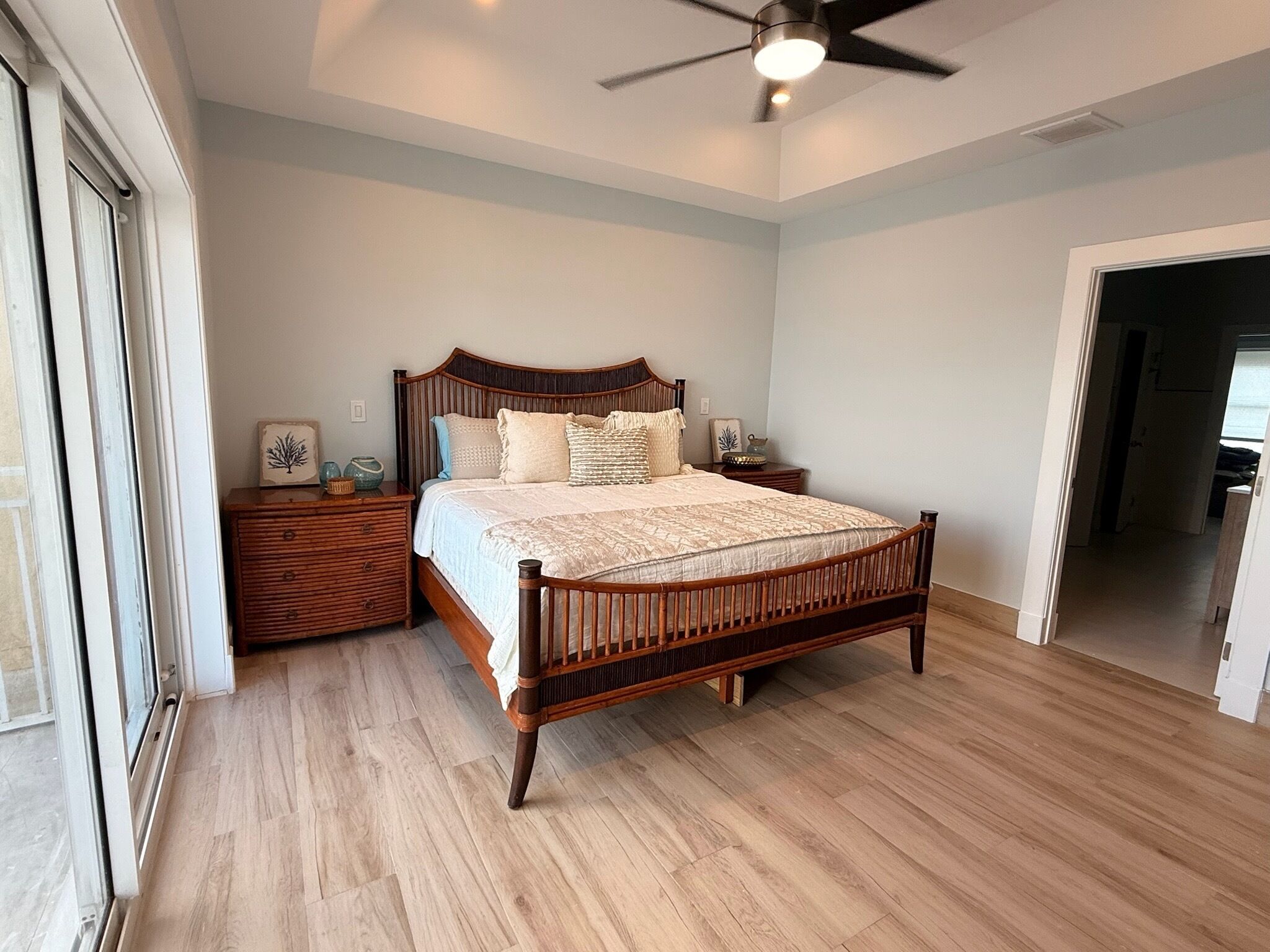 Un dormitorio con cama de madera, una mesita de noche, un ventilador de techo y vistas a través de un ventanal.
