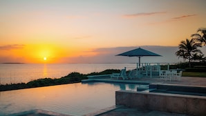 Una piscina infinita con vistas a la puesta de sol sobre el océano, sillas de descanso y un pabellón con sombrilla.