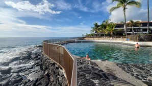A pool with a curved wooden barrier, a view of the ocean, and a building with a red roof in the background. - Kitchen and Bath Remodel! - Ocean Front - Central Air - Featured on Today Show (Kailua-Kona)