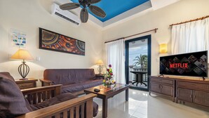 Una sala de estar con sofá, mesa de centro y televisor. Cuenta con ventilador de techo, aire acondicionado y un balcón con cortinas.