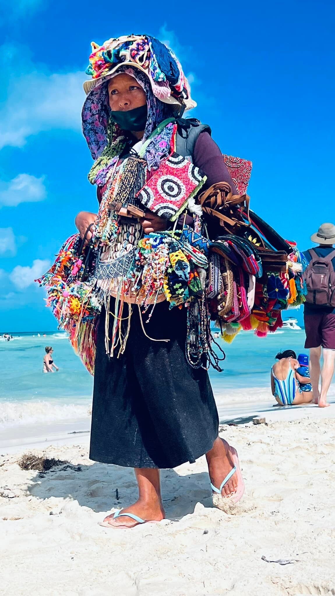 A person on a beach with a colorful, patterned outfit and a headpiece, carrying a large, decorated bag.