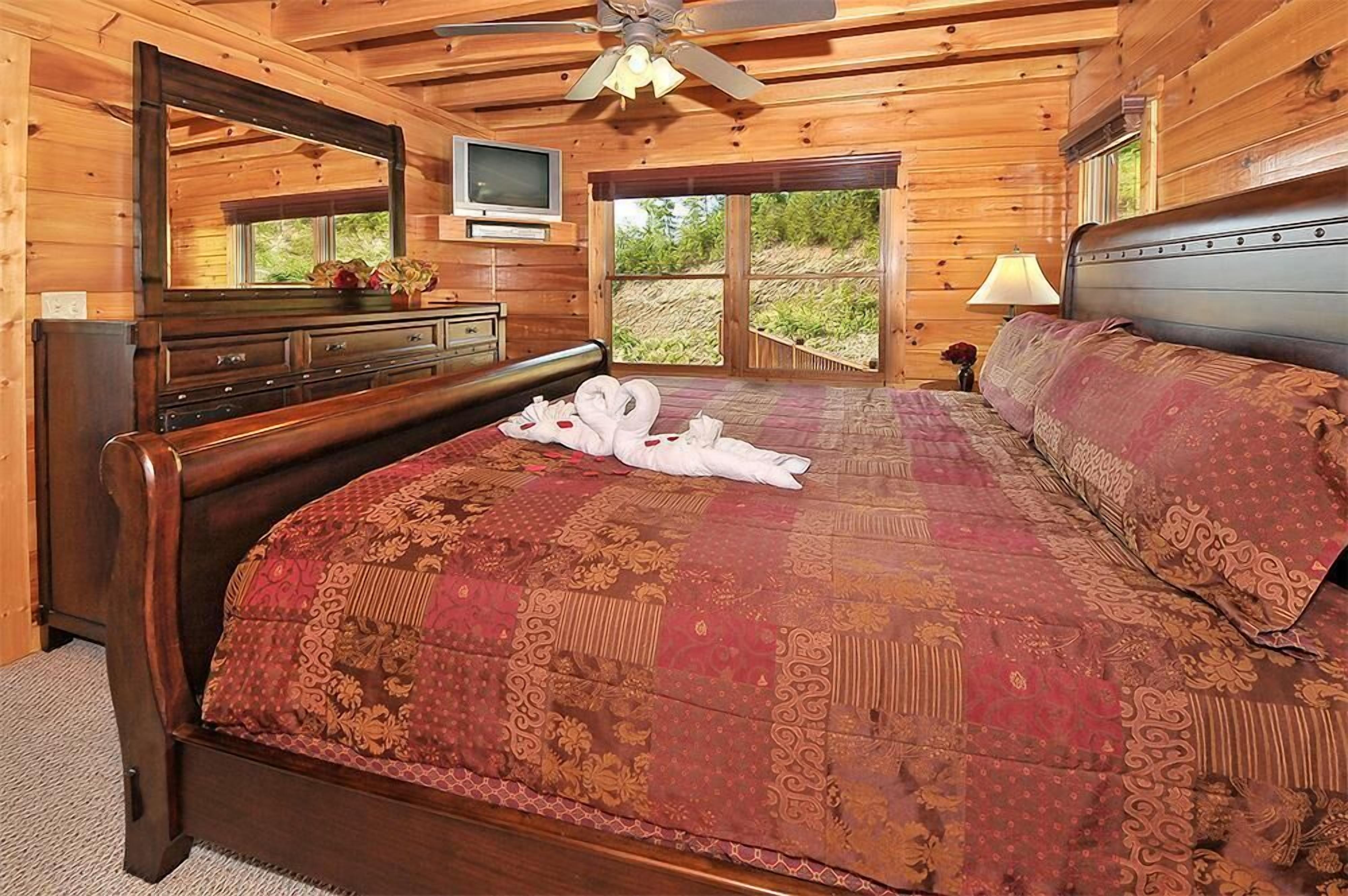 A wooden cabin bedroom with a large bed, a dresser, a TV, and a window with a view of greenery.