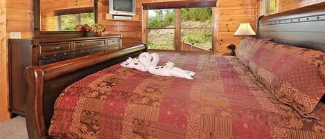 A wooden cabin bedroom with a large bed, a dresser, a TV, and a window with a view of greenery.