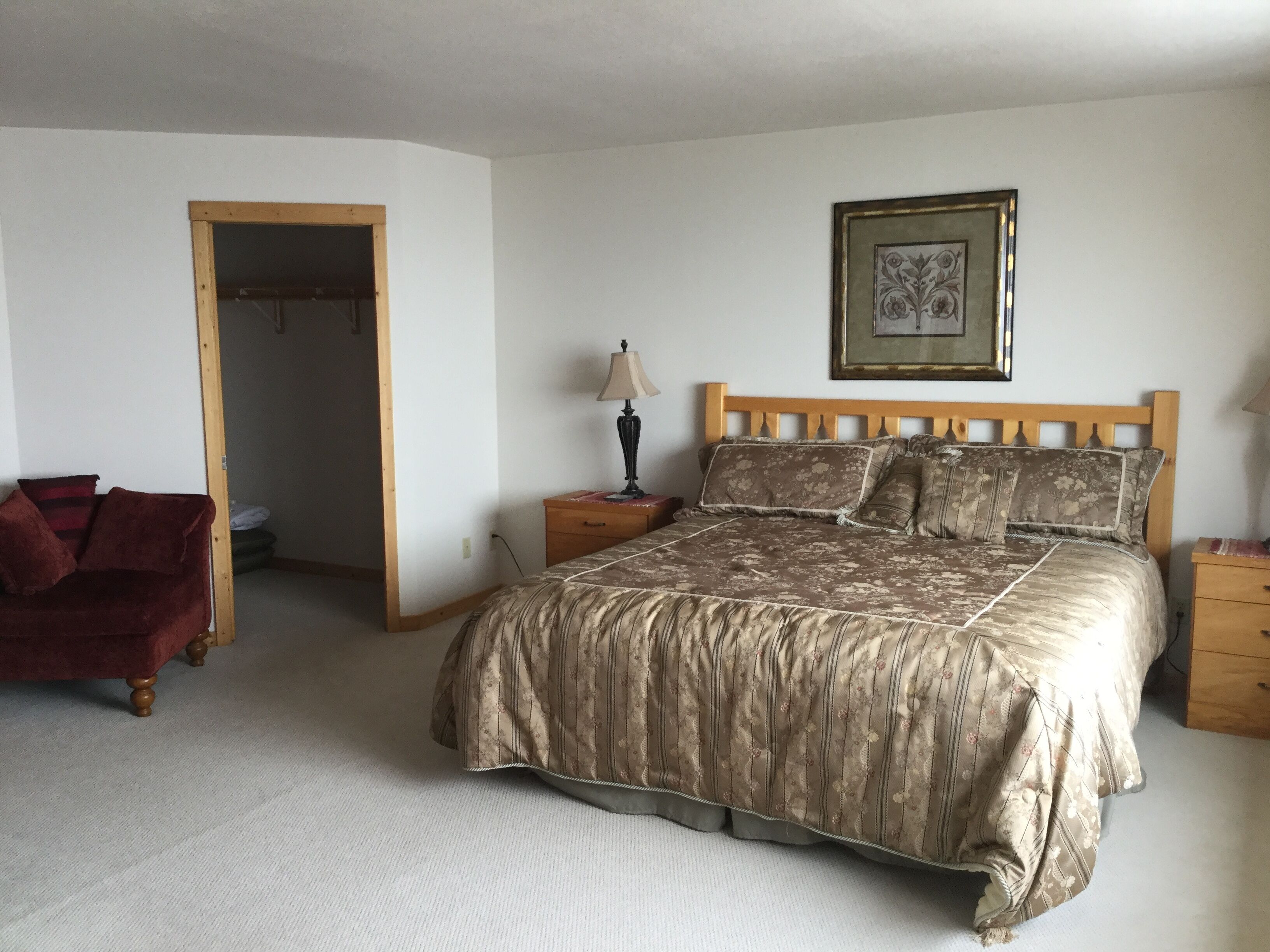 A bedroom with a large bed, a wooden headboard, a nightstand with a lamp, a framed picture on the wall, and a red chair.