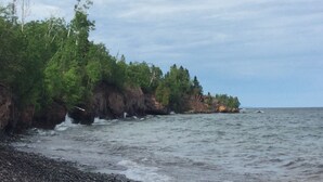 A rocky shoreline with waves crashing onto the pebbles, surrounded by dense green foliage and a clear sky.