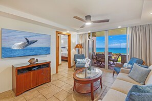 A spacious living area with a large TV displaying a whale, a comfortable sofa, a coffee table, and a view of the ocean through large windows. - Where Turtles and Sunsets Meet Kihei Oceanfront Condo Sweeping Ocean Views (Kihei)