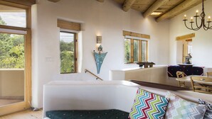 Een moderne kamer met een witte bank, een eettafel met stoelen, een kroonluchter en een groot raam met uitzicht op bomen.