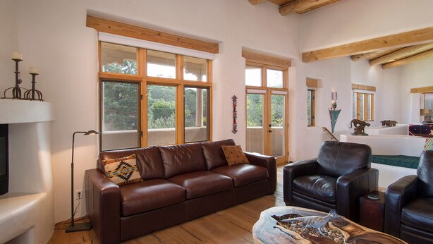 Een ruime woonkamer met een bruine leren bank, een zwarte leren fauteuil en een houten salontafel. Grote ramen zorgen voor natuurlijk licht en uitzicht op de omgeving.