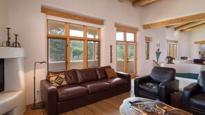 Un salon spacieux avec un canapé en cuir marron, un fauteuil en cuir noir et une table basse en bois. De grandes fenêtres laissent entrer la lumière naturelle et offrent une vue sur l’extérieur.