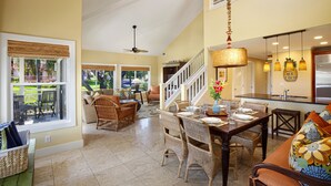 A spacious living area with a dining table, wicker furniture, and a staircase leading to an upper level. - Regency Villas 120 - Luxury 3BR Poipu Kai Condo | Pool, Lanais & Central AC (Koloa)