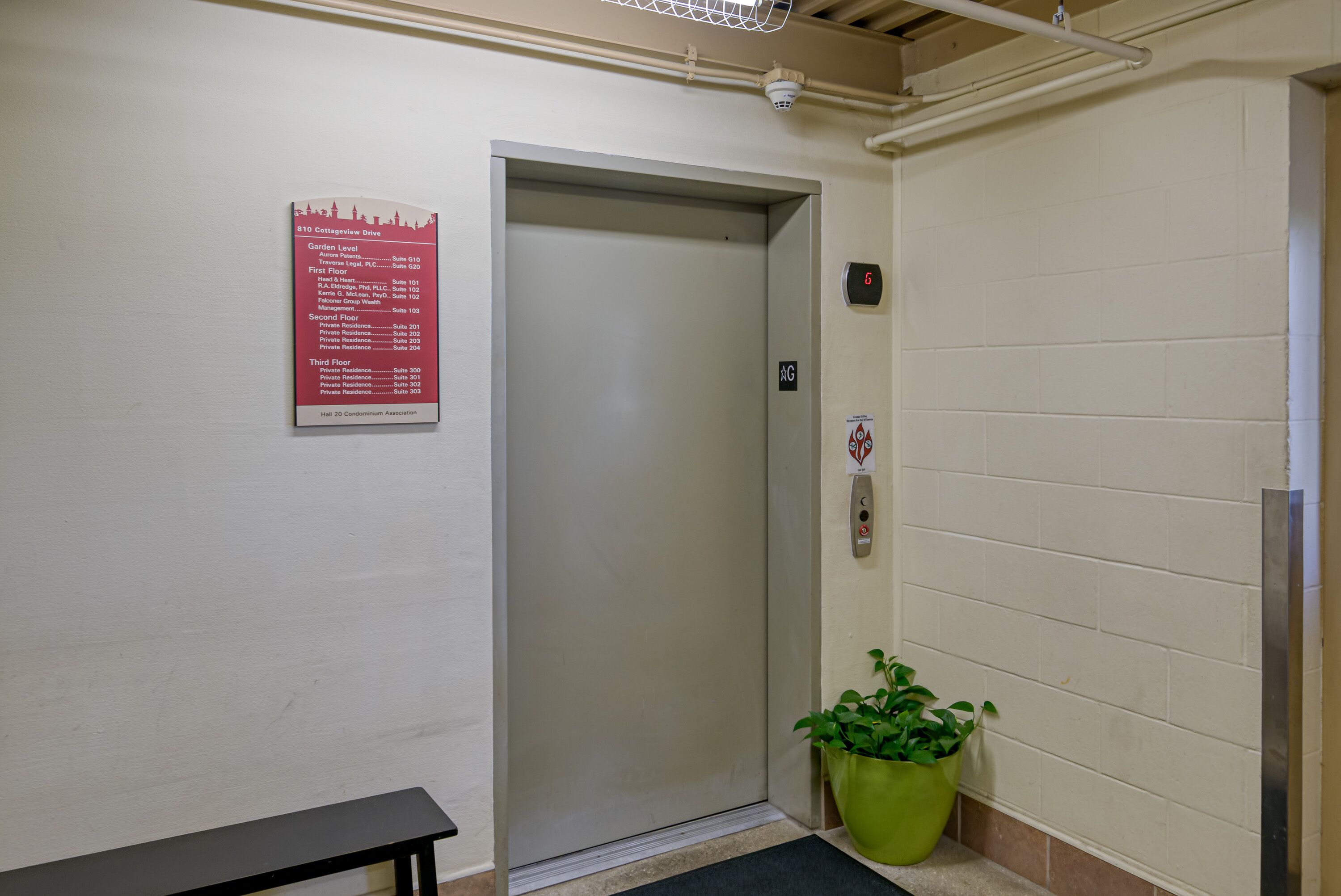 An elevator with a sign listing floor names and numbers, a green potted plant, and a bench.